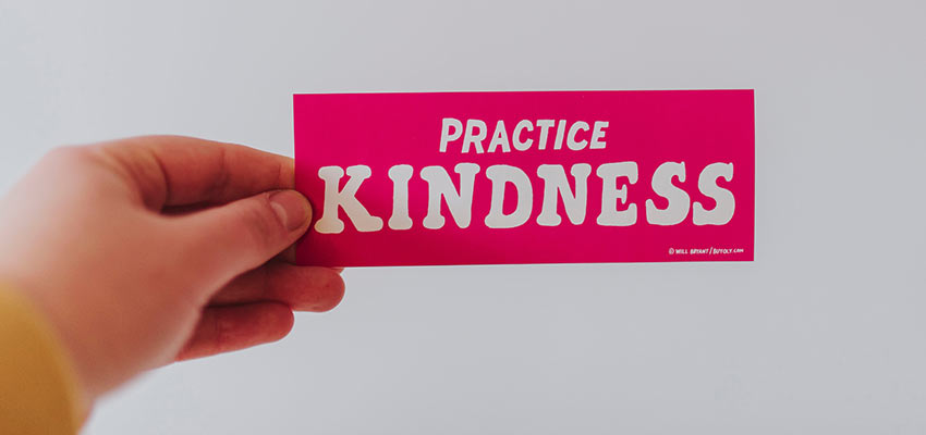 Person holding a bumper sticker that reads "Practice Kindness".